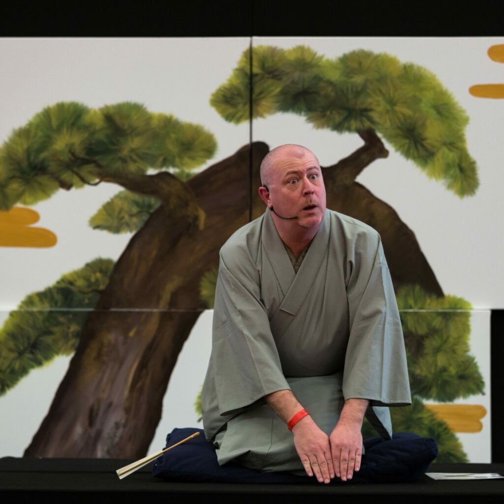 L’artiste Cyril Coppini de rakugo (stand-up) se tient au premier plan agenouillé sur un coussin, vêtu d’un kimono, il exprime sa surprise par les traits de son visage, derrière lui se trouve une peinture d’un arbre japonais. Le rakugo, du stand-up qui se joue assis, en kimono. Dimanche 7 avril, à 15h30, la salle des Éduens à Nevers, reçoit l’artiste Cyril Coppini.