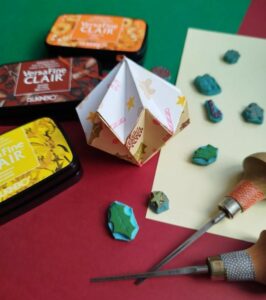 Mise en scène de linogravure avec de l'encre, des tampons et des gouges. On voit au centre la réalisation final : une suspension en origami décorée